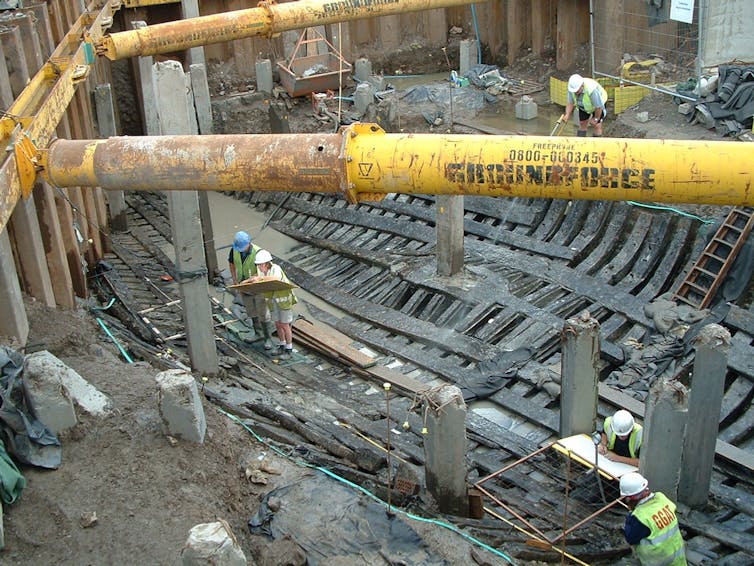 A group of people wearing hard hats and high-visibility vests stand within a construction site on timber planks