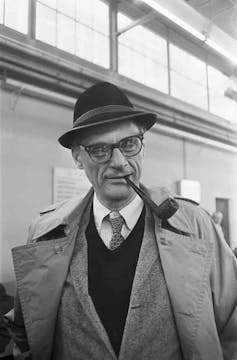 A man in a trench coat and hat looks at the camera while smoking a pipe.