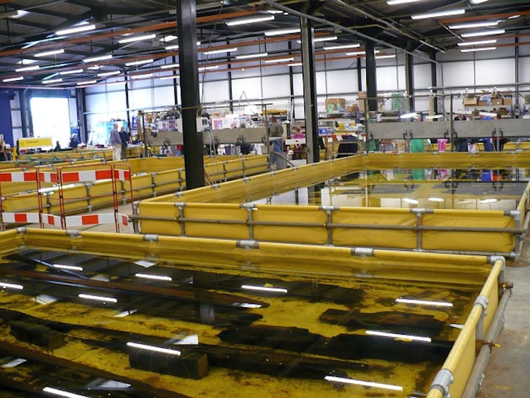 Wooden boards are in water in large but flat yellow baths in a large warehouse.