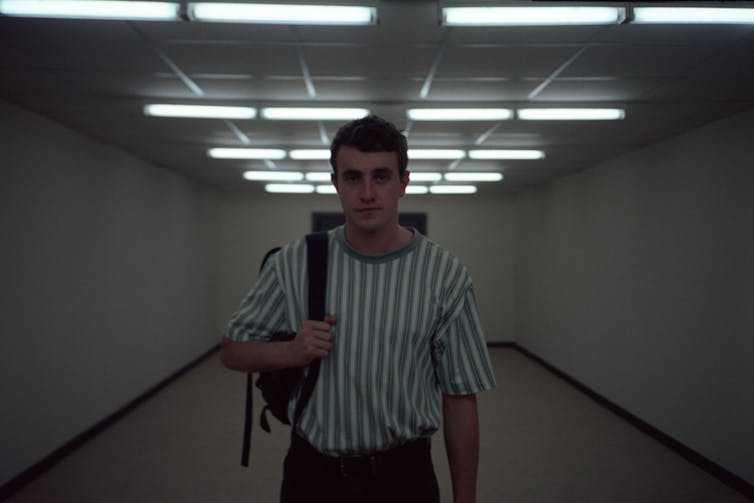 Paul Mescal stands in a wide white corridor wearing a striped t-shirt with a backpack strap over one arm. He stares straight ahead.