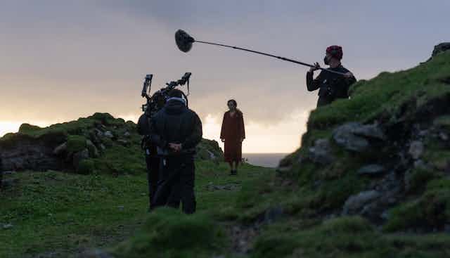 Kerry Condon stands between two grassy knoll wearing a red wool coat. In the foreground is a boom operator and camera man.