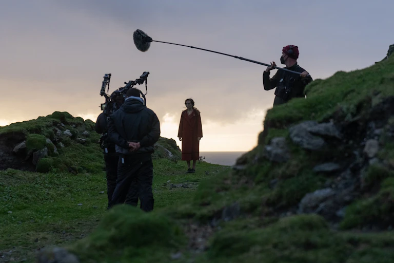 Kerry Condon stands between two grassy knoll wearing a red wool coat. In the foreground is a boom operator and camera man.