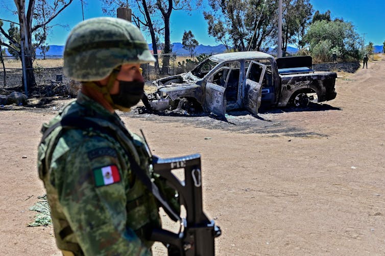 A soldier wearing camouflage and carrying a gun stands in front of a charred car on a sunny day.