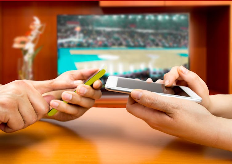 two pairs of hands hold smartphones with TV showing basketball game in background
