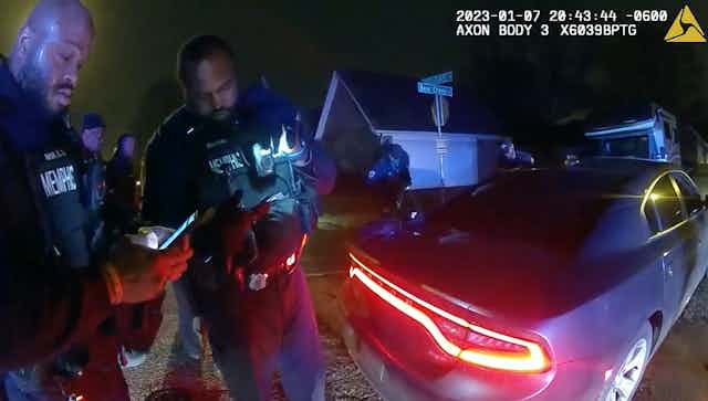 A video still shows two police officers in uniform stand next to a car.