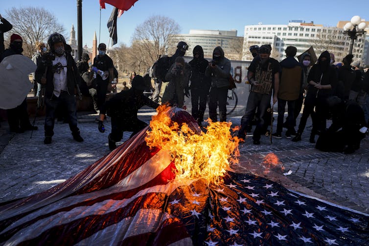 A group of people stand nearby while a U.S. flag burns.