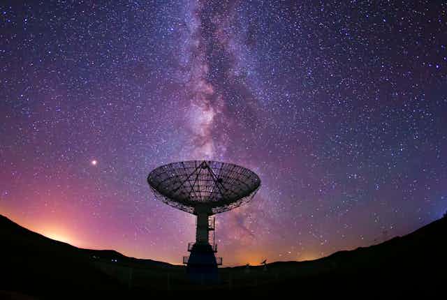 The semi-circular dish of a radio telescope silhouetted against a purple night sky with milky way directly vertical above it