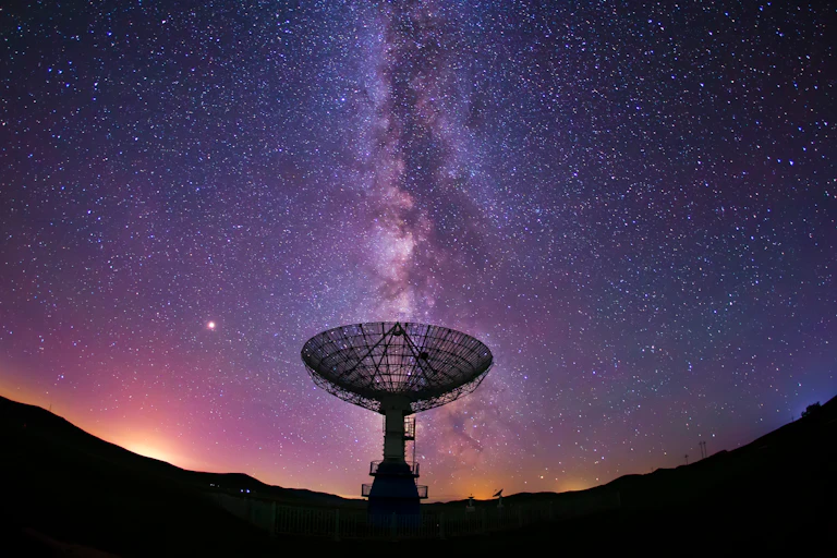 The semi-circular dish of a radio telescope silhouetted against a purple night sky with milky way directly vertical above it