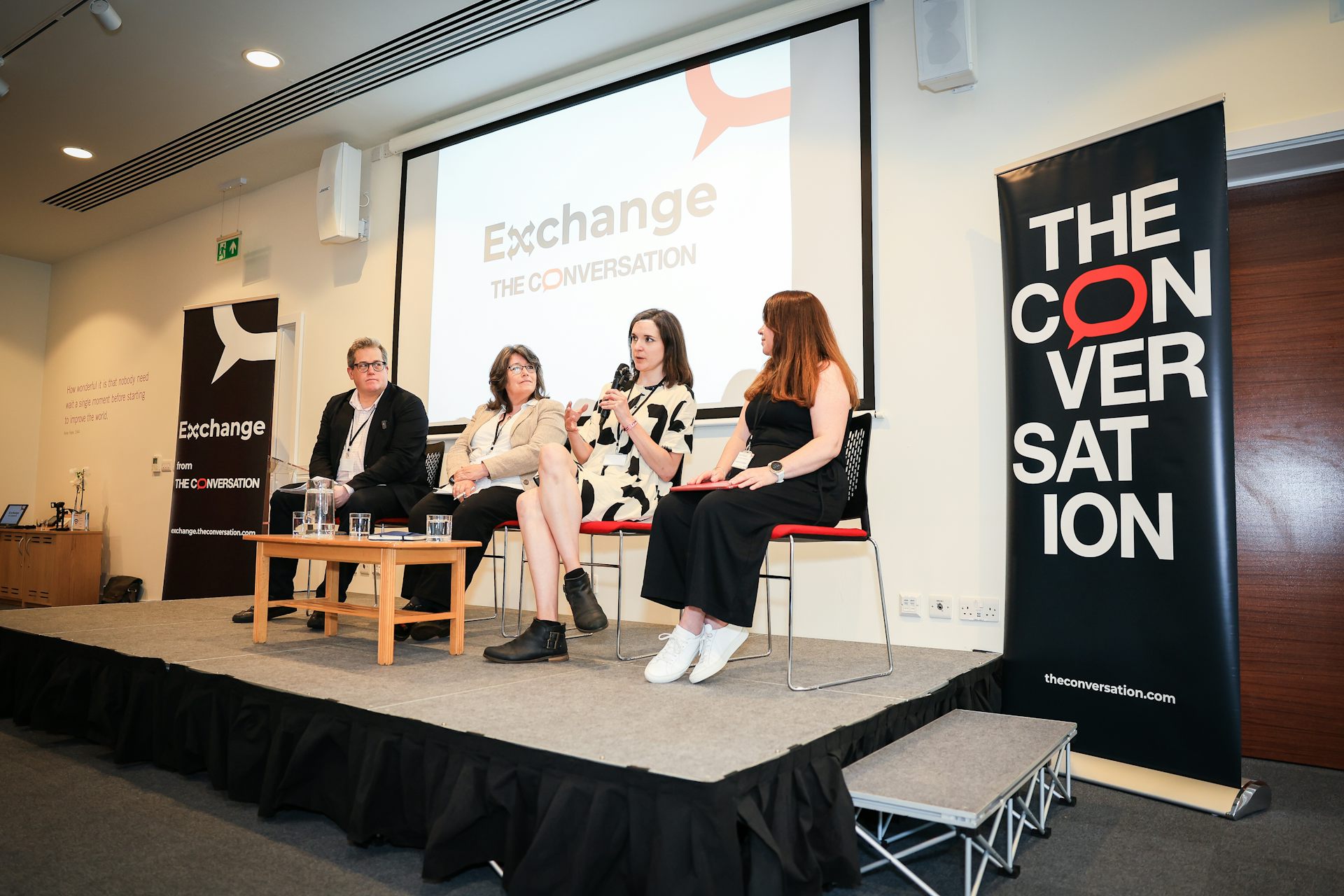 Panel members on a raised stage, flanked by branded banners bearing The Conversation's logo.