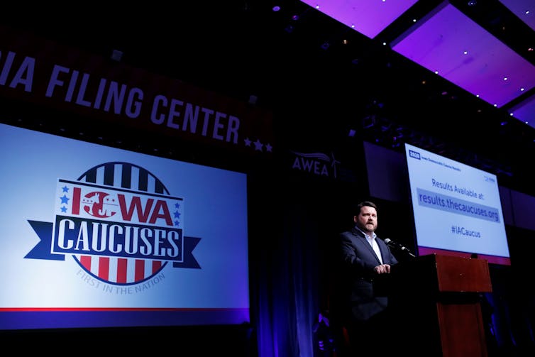 A suited man stands on stage, with his right hand on a lectern and speaking into a microphone. Behind him on his left is a screen with the message, Results Available at results.thecaucuses.org. And, to his right another screen bears the red, blue and white logo of the Iowa Caucuses.