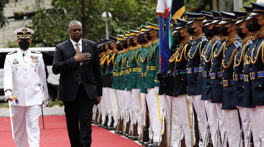 A middle aged Black man wearing a suit walks with his arm over his chest, next to a person with a mask in a white military suit. They walk past a row of soldiers all wearing blue and green and white pants.