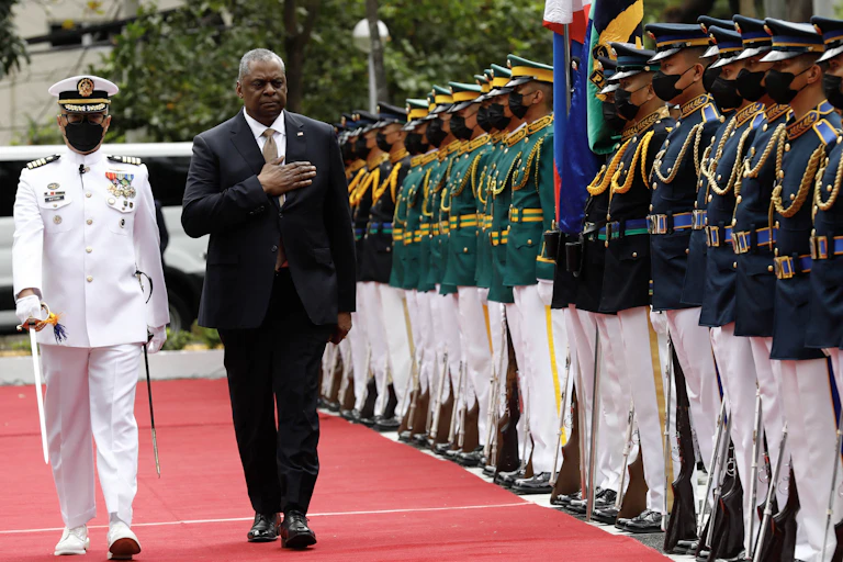 A middle aged Black man wearing a suit walks with his arm over his chest, next to a person with a mask in a white military suit. They walk past a row of soldiers all wearing blue and green and white pants.