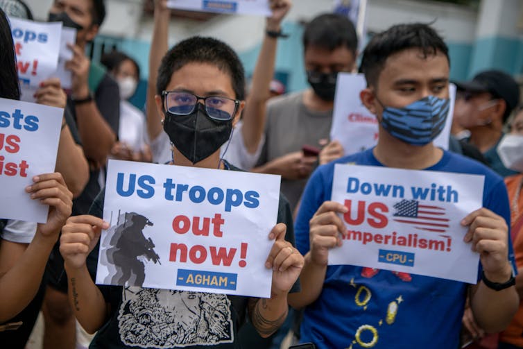 Two people hold up signs to their faces that say 'US troops out now' and 'Down with US imperialism.'