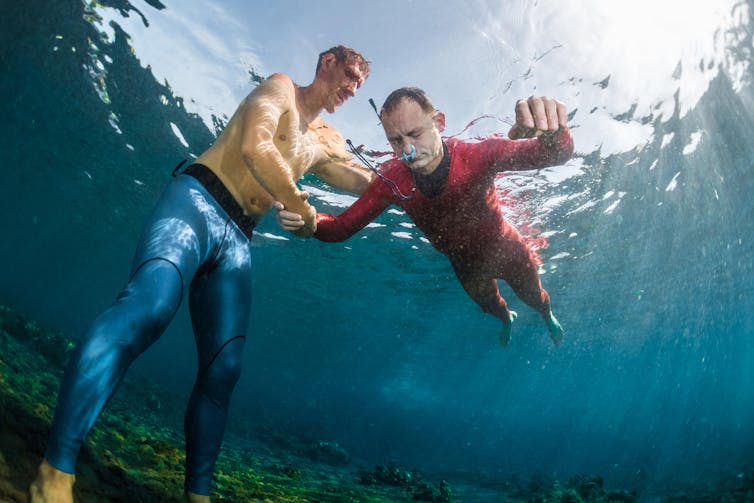 A man in a red wetsuit seen from underwater, holding his breath at the surface while a man in blue leggings assists him.