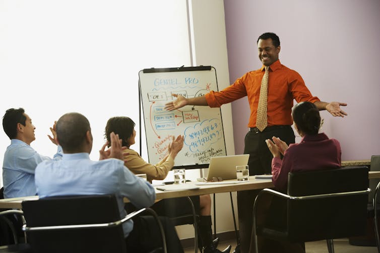 man stands soaking in applause from people around conference table