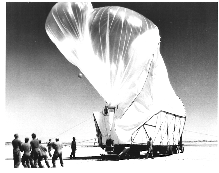 fotografía en blanco y negro de un grupo de hombres sujetando cuerdas atadas a un gran globo que está siendo inflado desde un camión en el desierto