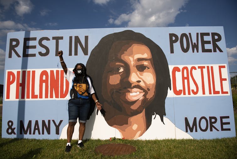 A black woman wearing a mask is standing next to large poster that has a portrait of her son. her son