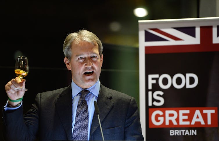 Owen Paterson holding up a glass of whiskey in front of a sign reading 'food is Great Britain'.