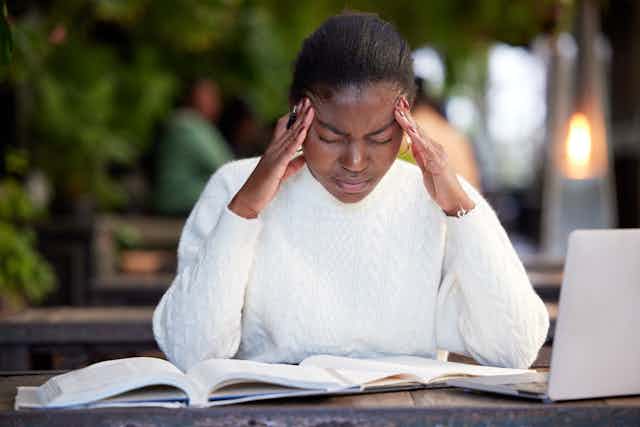 Young woman sitting at a table with books and a laptop, looking stressed with hands on head