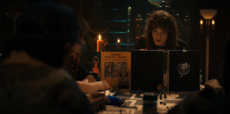 Eddie Munson sits behind the dungeon master's board. he has long curly hair and is lit by candles.