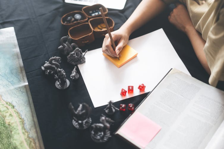 A hand writes on a sticky note next to the player's handbook.