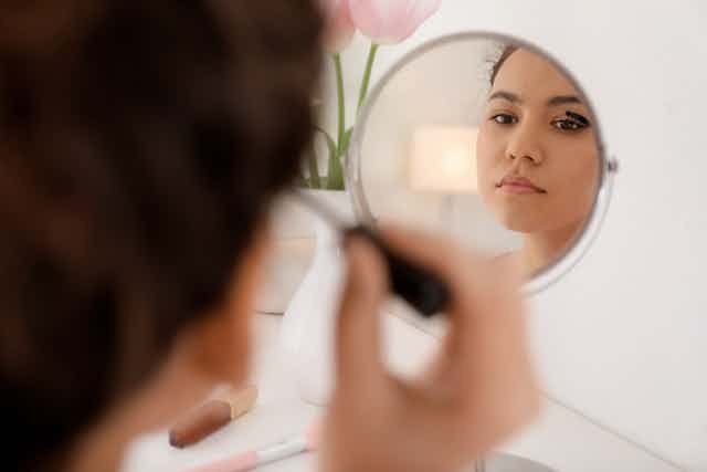 A young woman looks into a small countertop mirror while applying mascara