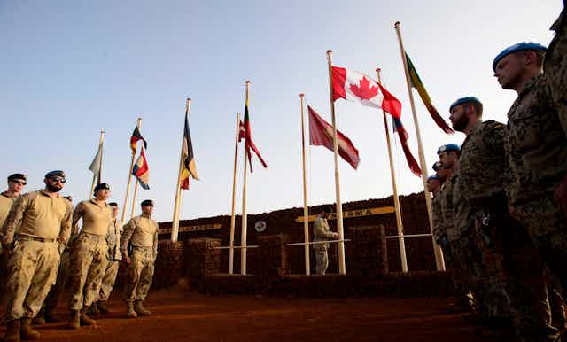 Soldiers in uniform stand at the foot of a row of flags, Canada's the most prominent.