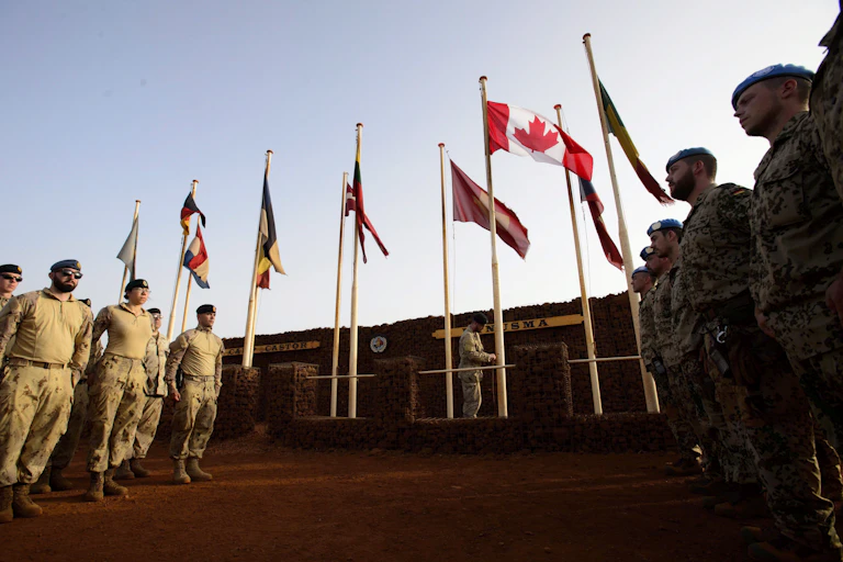 Soldiers in uniform stand at the foot of a row of flags, Canada's the most prominent.