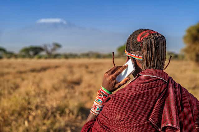 A woman in traditional African clothes is seen from behind staring across a savannah with Mount Kilimanjaro in at the background, talking on a cellular phone.