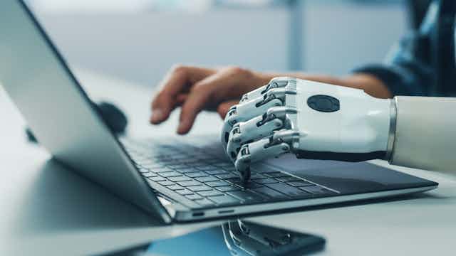 Una mano derecha humana y otra izquierda de robot escriben en el mismo teclado de un ordenador portátil.