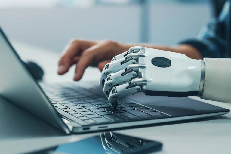 Una mano derecha humana y otra izquierda de robot escriben en el mismo teclado de un ordenador portátil.