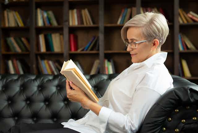 Una mujer de perfil con camisa blanca, pelo corto canoso y gafas lee un libro sentada en un sofá negro.