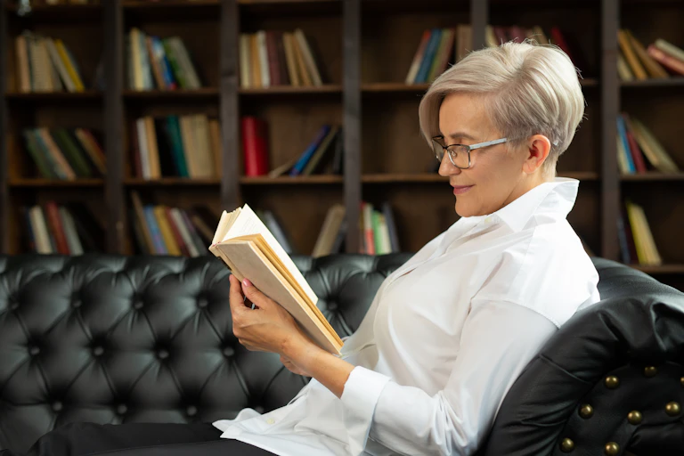 Una mujer de perfil con camisa blanca, pelo corto canoso y gafas lee un libro sentada en un sofá negro.