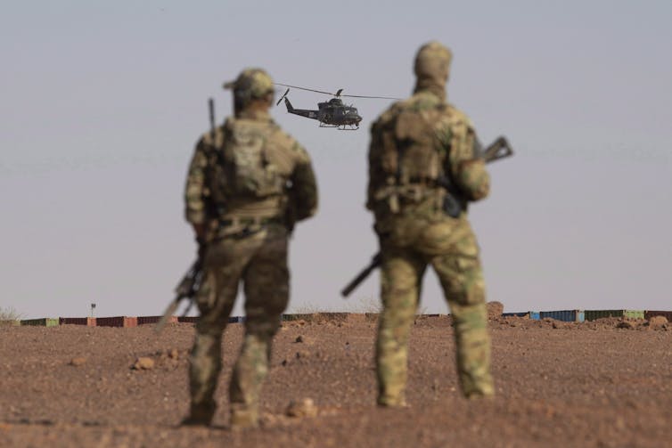 Two soldiers in battle fatigues stand in a dusty field and watch as a helicopter approaches.