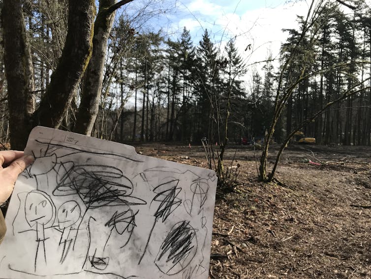 A hand is seen holding a child's drawing in front of woods.