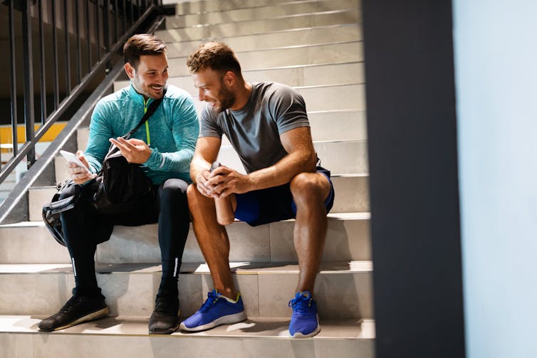 Two young men wearing workout clothes sit on a staircase and talk.