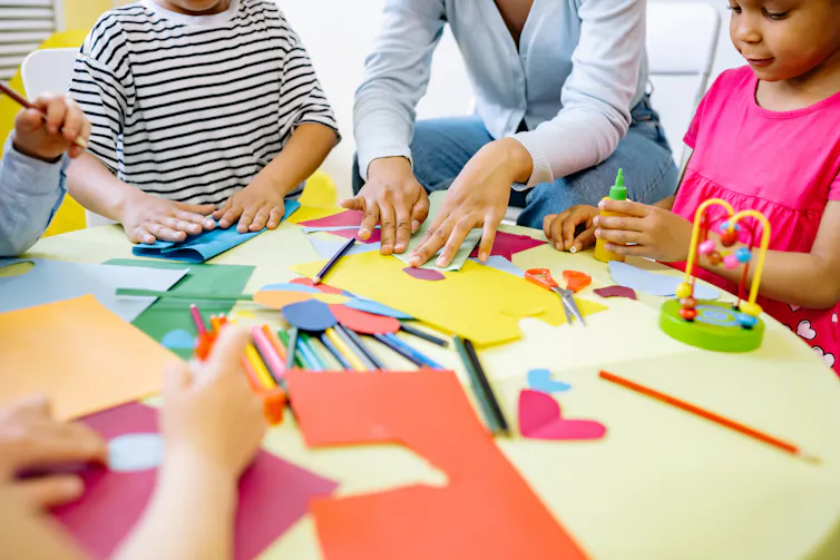 Children's and an educator's hands seen working with paper at a table.