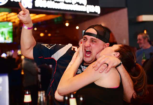 Excited man in football jersey and backwards hat being kissed by a woman on the cheek.