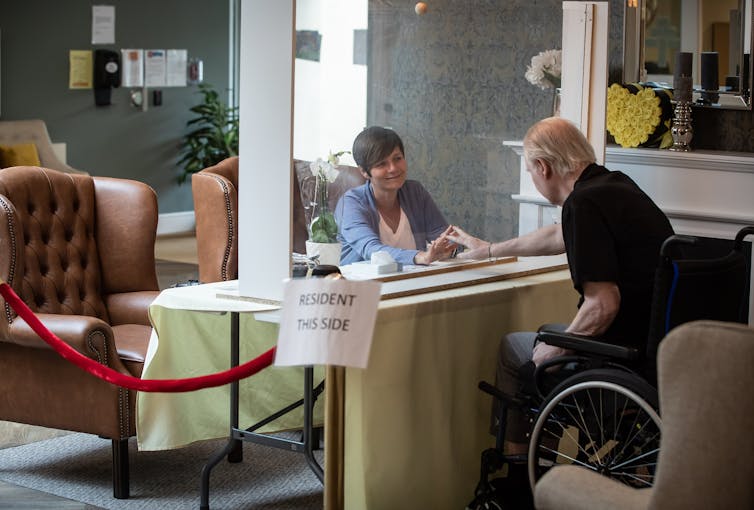A younger woman and an elderly man in a wheelchair place their hands on a glass barrier separating them.
