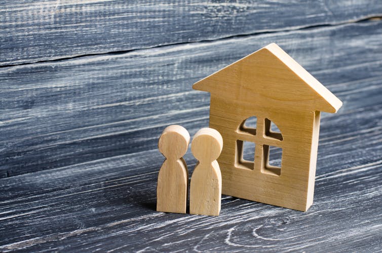 Two wooden figures are set in front of a small wooden house.