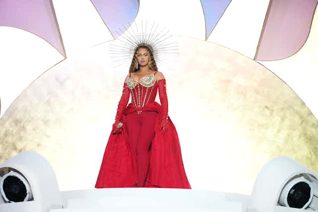 A woman stands on a stage wearing a red dress with a bustle and a crown on her head.