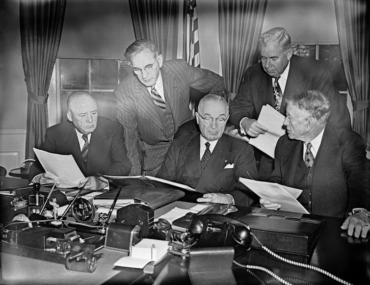 Five men - three sitting, two standing - in suits looking at pieces of paper at a desk.