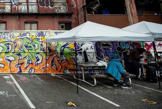 Picnic tables under temporary shade structures in a parking lot in front of a wall covered in graffiti; a person in a blue jacked is slumped over at one of the tables