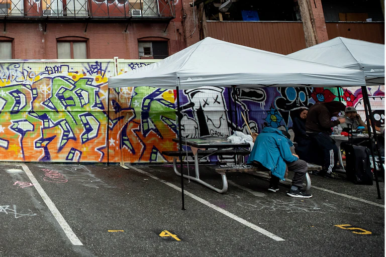 Picnic tables under temporary shade structures in a parking lot in front of a wall covered in graffiti; a person in a blue jacked is slumped over at one of the tables