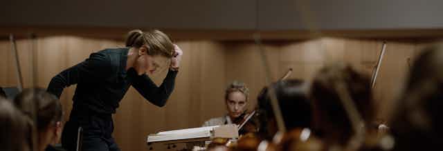 a white woman, standing, head down, fist in the air, conducts an orchestra