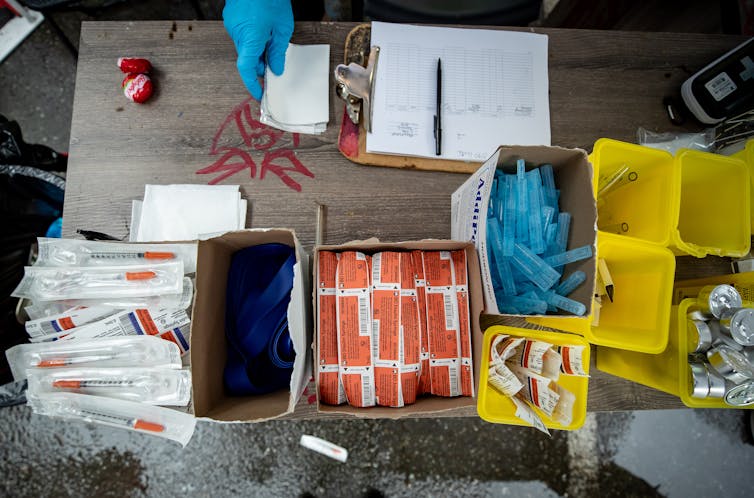 Boxes of syringes and bins of glass tubes, candles and matches on a table with a clipboard. A gloved hand is seen at the top of the photo.