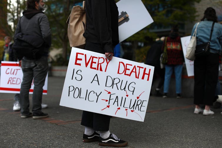 Cropped image of a person holding a hand-lettered sign reading 'Every death is a drug policy failure'