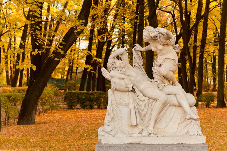 A statue of a naked woman looking down at a sleeping man on display in a park in autumn.