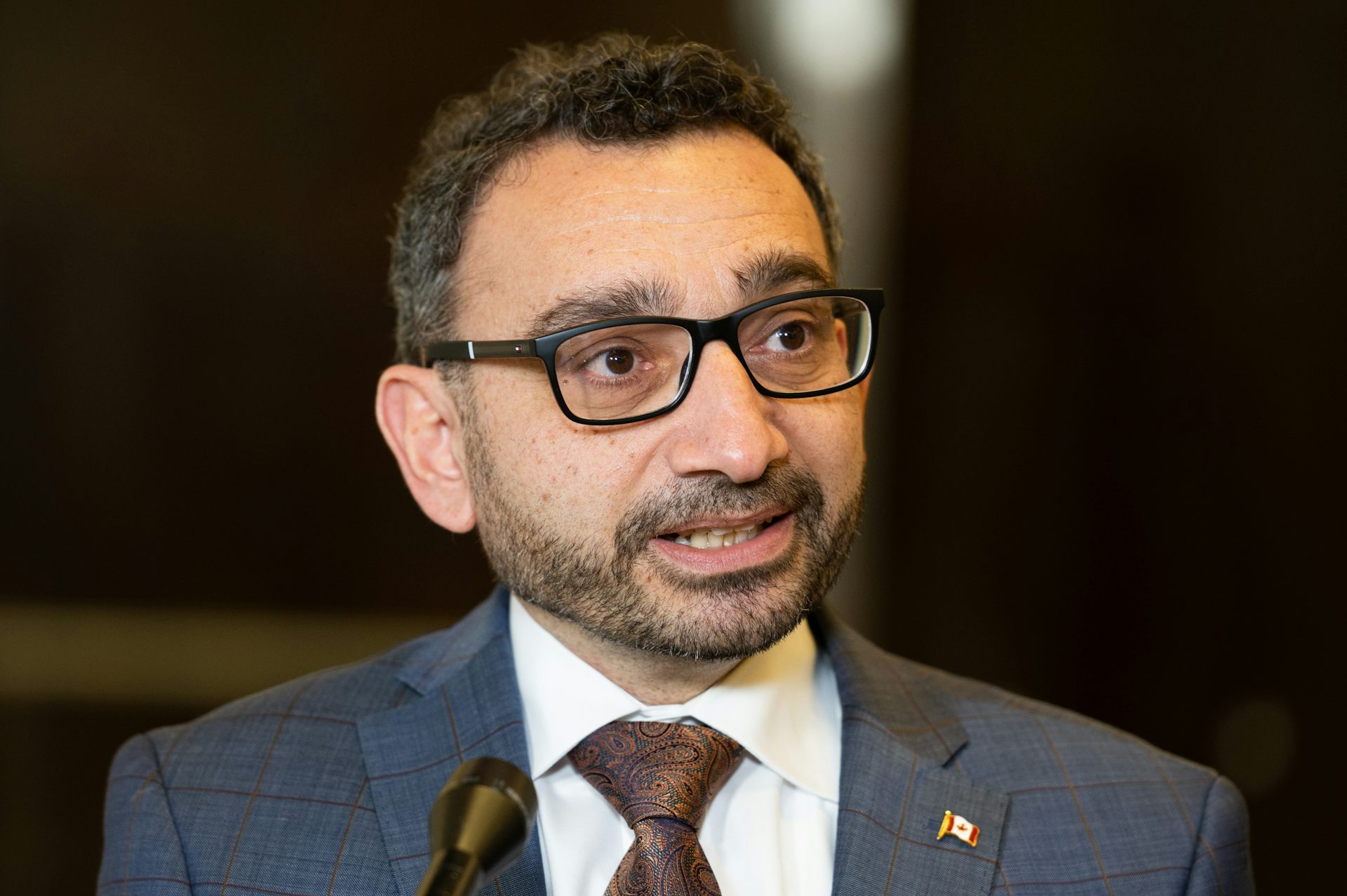 A man in a suit and tie wearing square glasses looks off-camera as he speaks