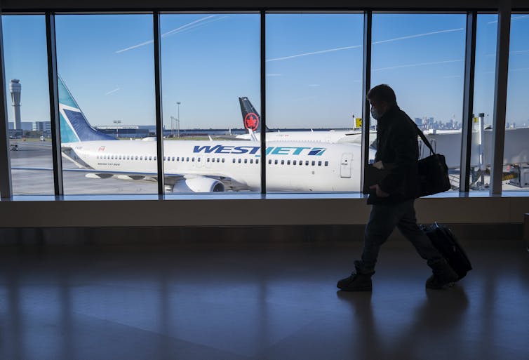 The silhouette of a man walks in front of a massive window overlooking an airport tarmac with a WestJet plane sitting on it
